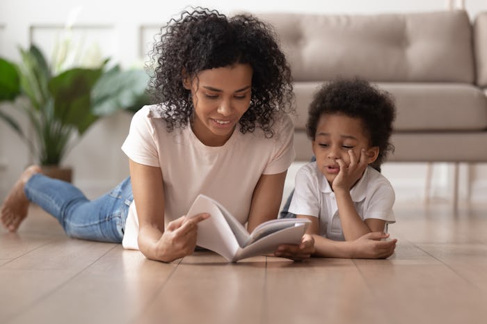 Young african American mom lying on floor with cute toddler boy reading book at home together, black...