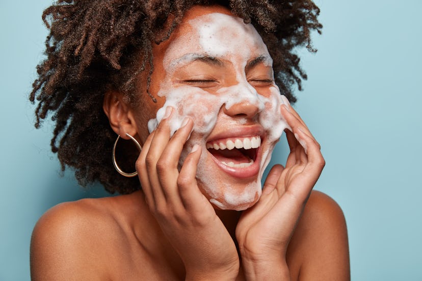 Cheerful black female model applies foaming cleanser, has clean fresh healthy skin, smiles broadly, ...