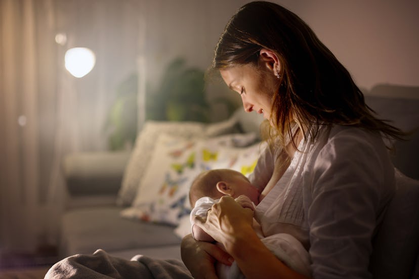 Young beautiful mother, breastfeeding her newborn baby boy at night, dim light. Mom breastfeeding in...