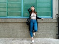 A woman leans against a wall, while wearing a white crop top and jeans. 