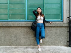 A woman leans against a wall, while wearing a white crop top and jeans. 