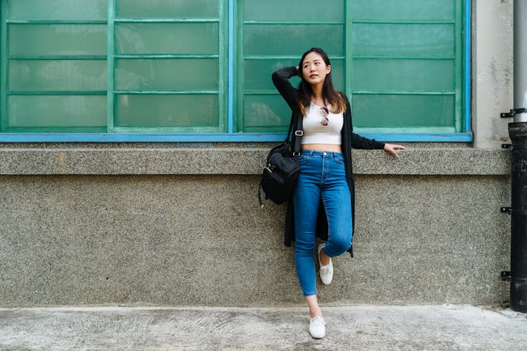 A woman leans against a wall, while wearing a white crop top and jeans. 