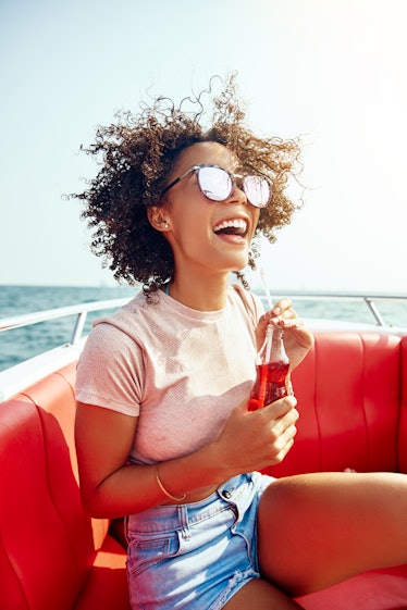 Laughing young woman wearing sunglasses and enjoying a drink while standing on a boat during summer ...