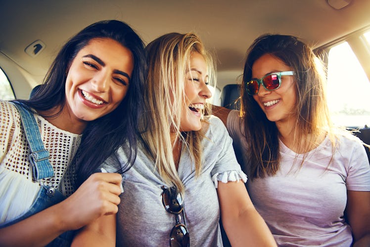 Laughing affectionate female friends riding in the back of a car on a road trip together smiling hap...