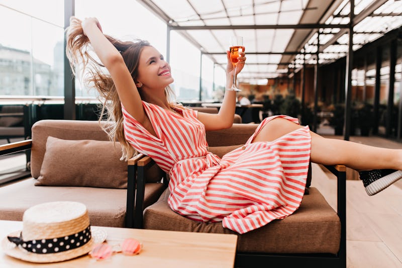 Ecstatic blonde girl playing with her hair while chilling in cafe. Inspired young woman in striped dress enjoying champagne in restaurant in summer weekend.
