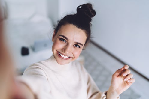 Pretty young female with big smile standing at bedroom after work with laptop and having fun taking ...