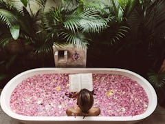 A young woman reads a book while soaking in a flower bath in the jungle.