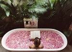 A young woman reads a book while soaking in a flower bath in the jungle.