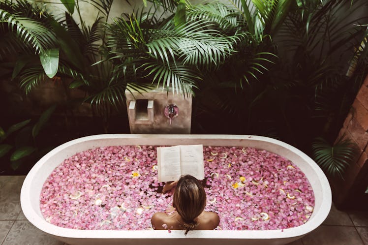 A young woman reads a book while soaking in a flower bath in the jungle.