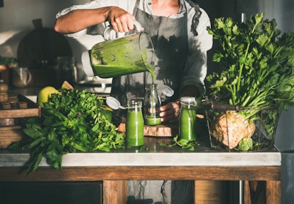 Making green detox take-away smoothie. Woman in linen apron pouring green smoothie drink from blende...