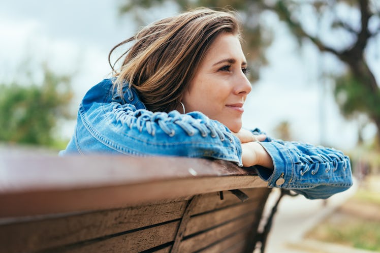 Young woman sitting on a bench outdoors in a park leaning on the back staring into the distance dayd...