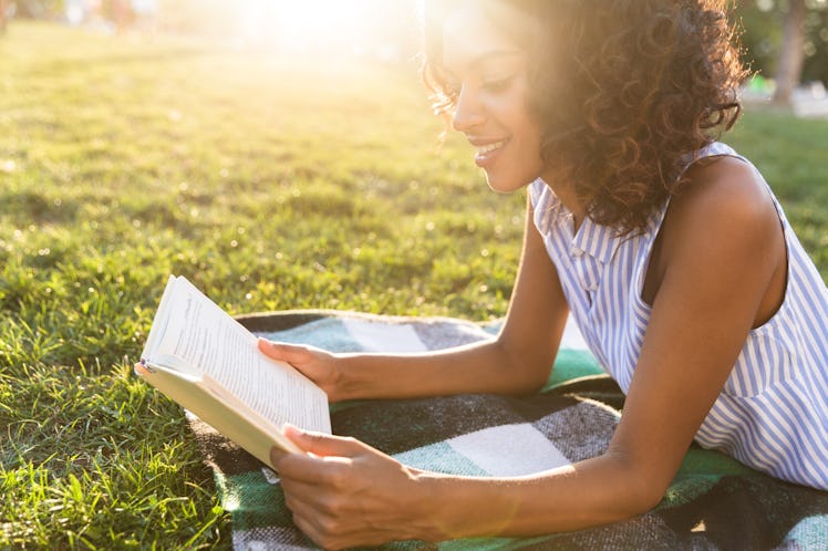 Happy african girl laying on a grass at the park, reading book
