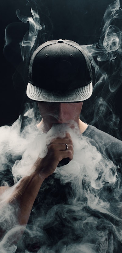 Vape man. Portrait of a handsome young white guy in a modern black cap vaping and letting off puffs ...