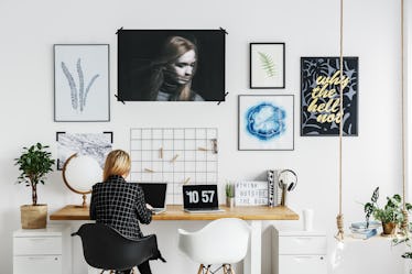 A blonde woman with a black and white flannel sits at a trendy desk in her home.