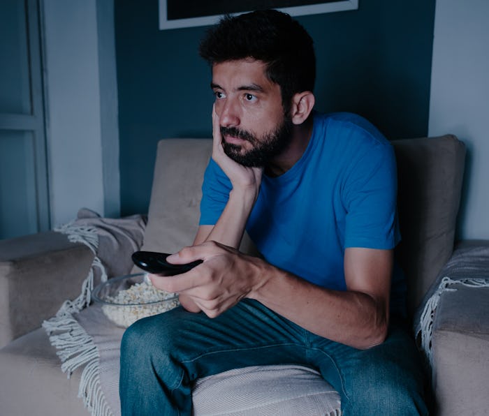 A man with prominent facial hair can be seen watching TV with a bored expression. His hand is cuppin...