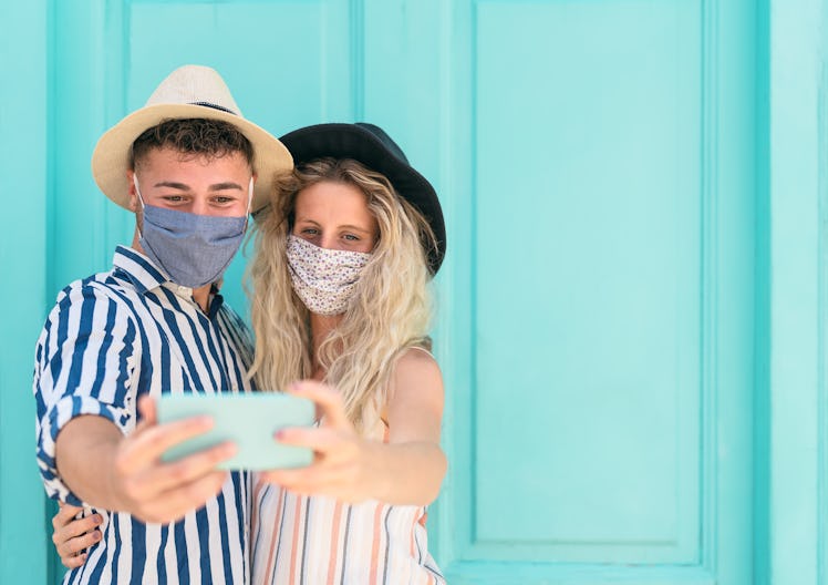 Young couple wearing face mask taking selfie with mobile smartphone on vacation - People having fun ...