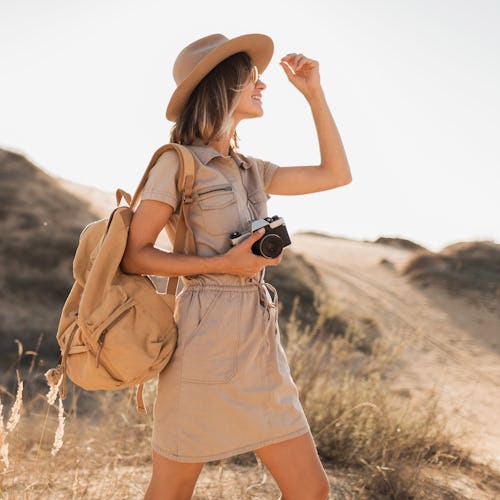 stylish young woman in khaki dress walking in desert, traveling in Africa on safari, wearing hat and...