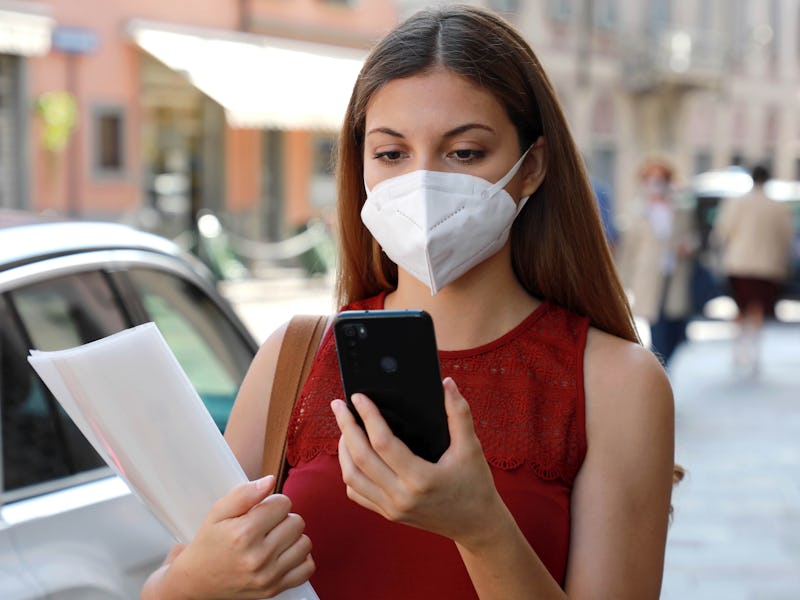A young woman wearing a face mask is seen holding a smartphone in one hand and papers in her other a...