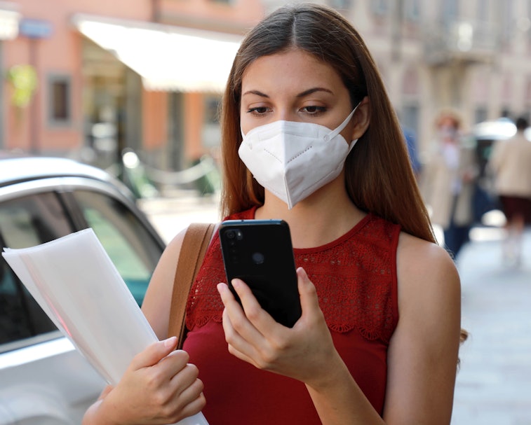 A young woman wearing a face mask is seen holding a smartphone in one hand and papers in her other a...