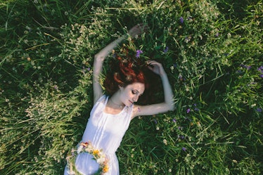 In spring, a young red-haired woman with a floral wreath  and in a white dress laying on a field on ...
