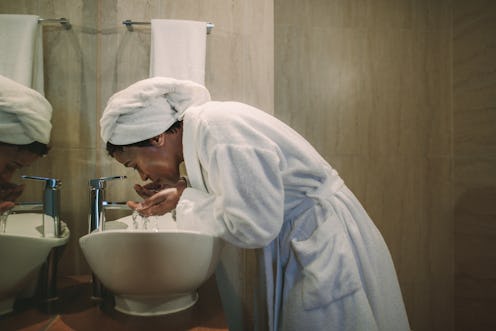 African woman in bathrobe washing her face in bathroom sink. Mature female washing her face in bathr...