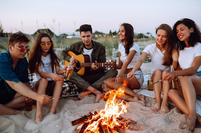 A group of friends hang out at the beach by a bonfire while roasting hot dogs and one plays the guit...