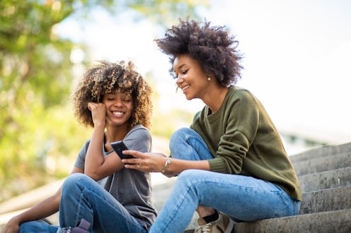 Side portrait two young african american women sitting on steps outside looking at mobile phone