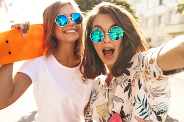 Two trendy girls smile and wear blue round sunglasses while posing for a selfie outside on a sunny s...