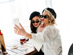 Two elegant women dressed in white button-down shirts, black hats, and sunglasses pose for a selfie ...