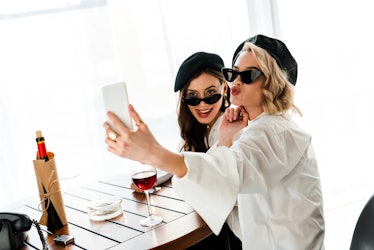 Two elegant women dressed in white button-down shirts, black hats, and sunglasses pose for a selfie ...