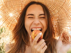 A happy woman wearing a sun hat bites into a peach in a field with the sun behind her.