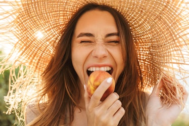 A happy woman wearing a sun hat bites into a peach in a field with the sun behind her.