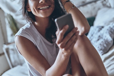 A woman smiles while cuddling on her bed and looking at her phone.