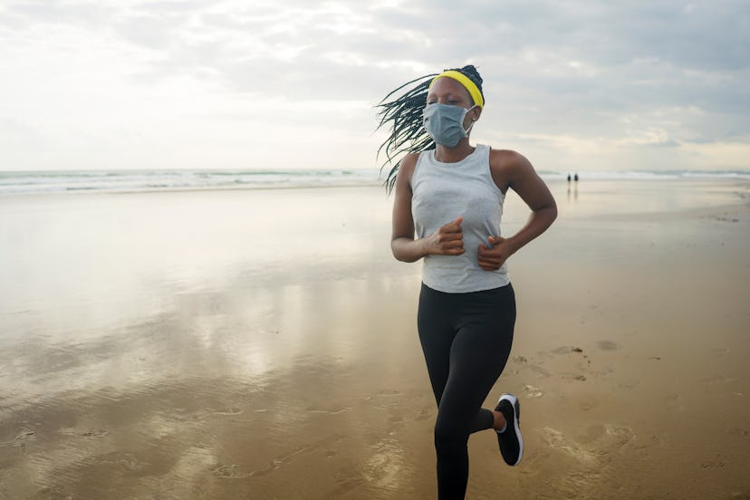 A person wearing a yellow headband and white tank top runs on the beach while wearing a mask. Going ...
