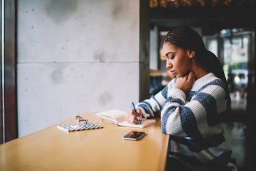 Thoughtful female student writing essay in textbook for education sitting in cafeteria, concentrated...