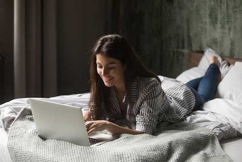 Smiling attractive woman lying on bed using laptop communicating online at home, happy girl typing o...