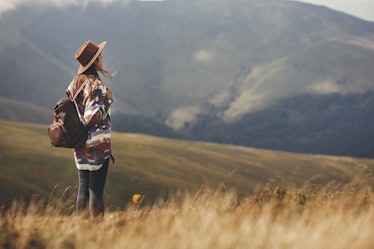 Stylish hipster girl in hat walking on top of mountains. Happy young woman with backpack exploring s...