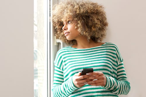 A woman is holding her phone and pensively looking out a window. Venus retrograde 2021 will affect C...