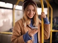 A happy woman wearing a beanie and jacket smiles while she texts on the subway.