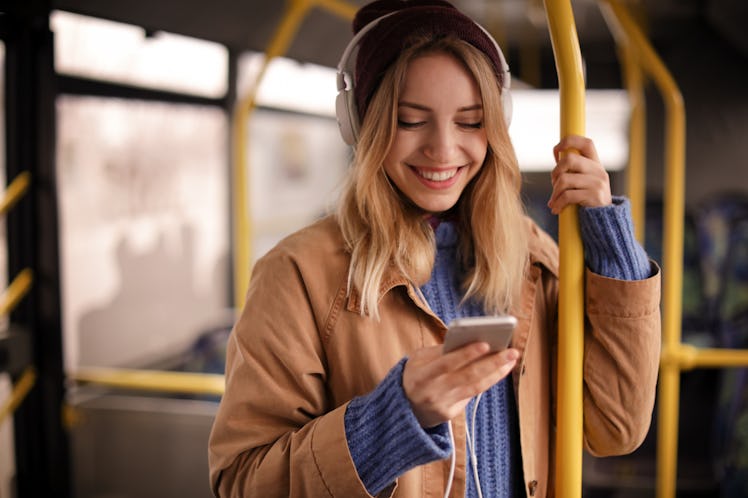 A happy woman wearing a beanie and jacket smiles while she texts on the subway.