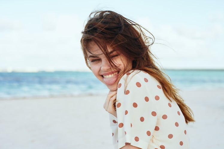 A happy woman wearing a white and brown polka dot shirt smiles while at the beach.