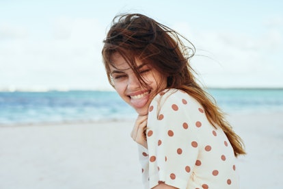 A happy woman wearing a white and brown polka dot shirt smiles while at the beach.