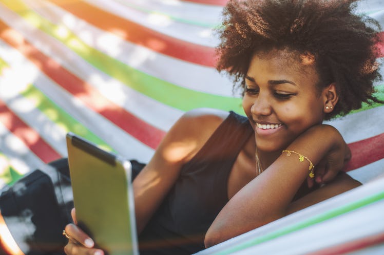 A happy woman reads a book on her tablet while relaxing in a hammock in the sunshine at the beach.