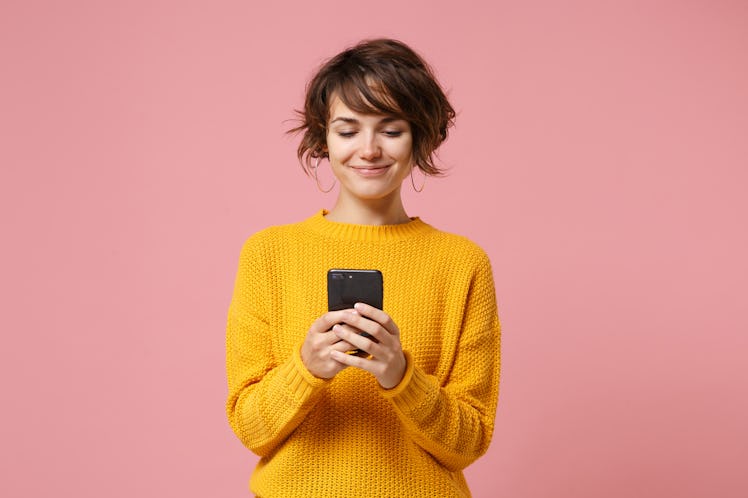 Smiling young brunette woman girl in yellow sweater posing isolated on pastel pink wall background s...