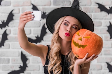 Beautiful blonde girl in black dress and witch hat is holding a scary pumpkin and doing selfie using...
