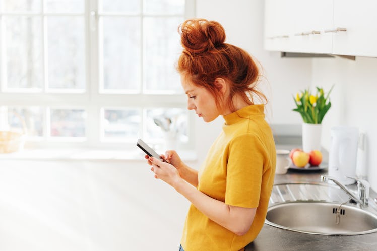 Young red-haired woman in bright yellow sweater standing and using smartphone, leaning on kitchen si...