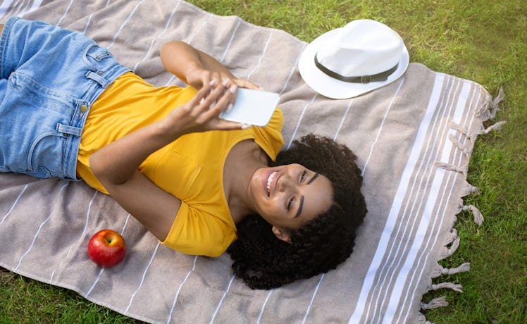 Lovely black woman using mobile phone on picnic blanket outside, top view