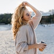 Stylish beautiful woman blonde in beige oversized sweater and brown sunglasses walks along beach wit...
