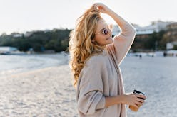 Stylish beautiful woman blonde in beige oversized sweater and brown sunglasses walks along beach wit...