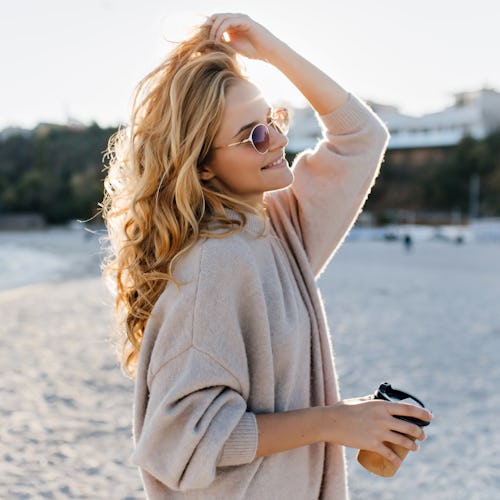 Stylish beautiful woman blonde in beige oversized sweater and brown sunglasses walks along beach wit...